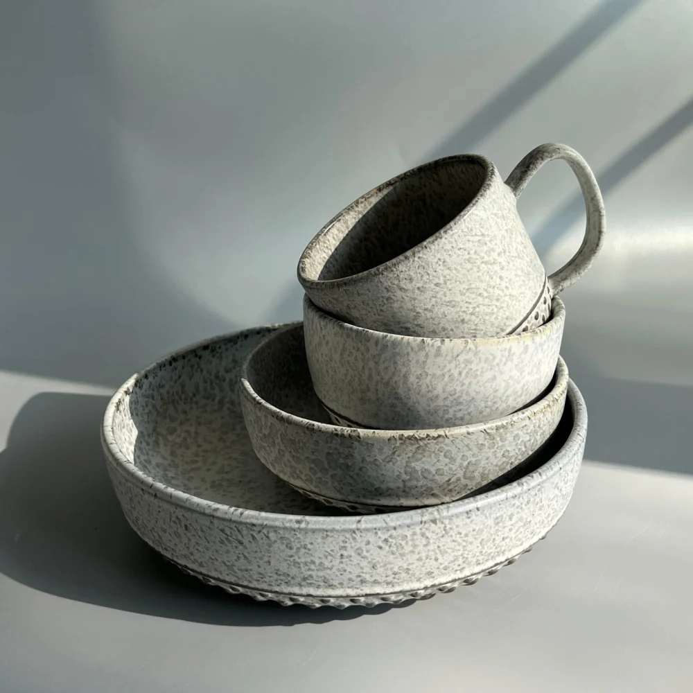 Official detail photograph of a pale speckled mug tilted over stacked bowls